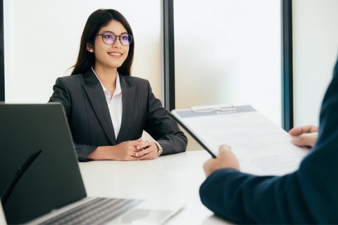 a young woman with dark hair, wearing a dark blazer and glasses, smiling at a person whose arm is visible in the foreground and is sitting across the table, holding a clipboard
