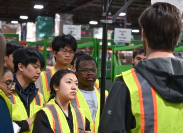 Group of students on warehouse floor wearing yellow safety vest looking at a staff engineer.