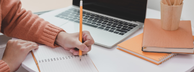 a person writing in a notebook with a pencil, next to a laptop computer
