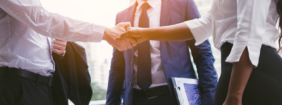 A woman shakes hands with a man in business apparel. A man in a suit stands in the background
