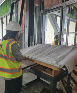 Inside of an in-progress building, Breanna stands thoughtfully in front of a large blueprint. She's wearing a bright construction vest and a hard hat.