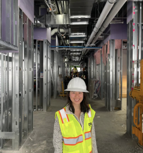 Breanna poses, smiling wide, in her construction vest and hard hat in the center of a hallway, the building in-progress around her.
