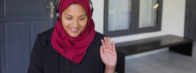 A woman wearing a hijab waves while on a virtual call.