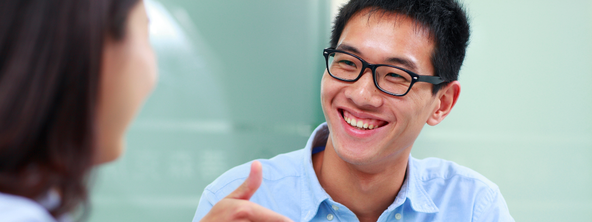A smiling man in glasses listens to another person.