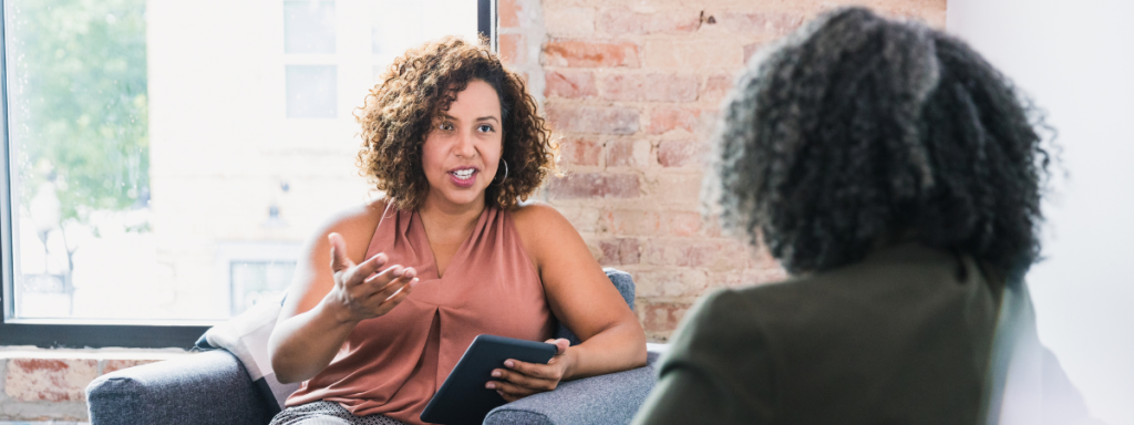 A woman holding a tablet advises another person in a comfortable conversation.