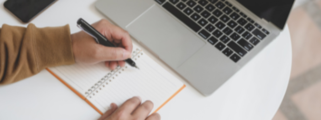 From above, a view of a person's hands as they write in a notebook. A laptop also sits on the table.