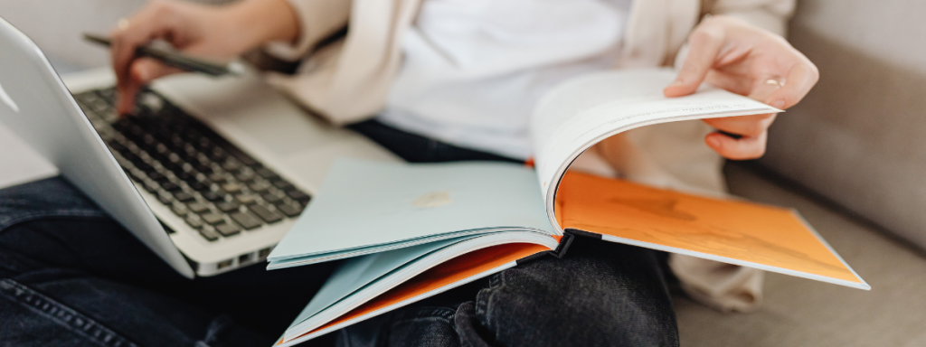 A person types on their laptop while looking at a book.