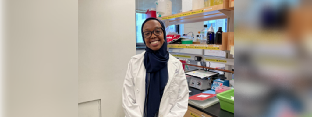 A student in a white coat poses, smiling, in the lab.