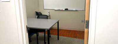 A view into a room from the outside hallway, featuring a table, chairs, and a whiteboard.